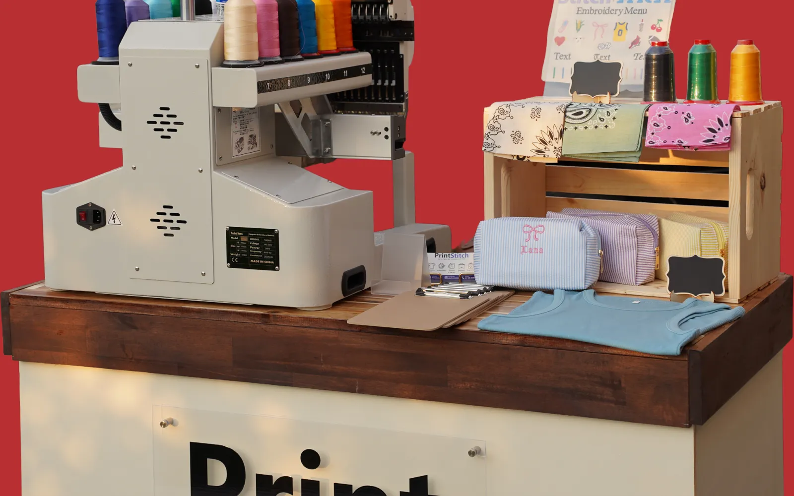 Close-up of the PrintStitch live embroidery cart: thread spools, a commercial embroidery machine, and monogrammed pieces