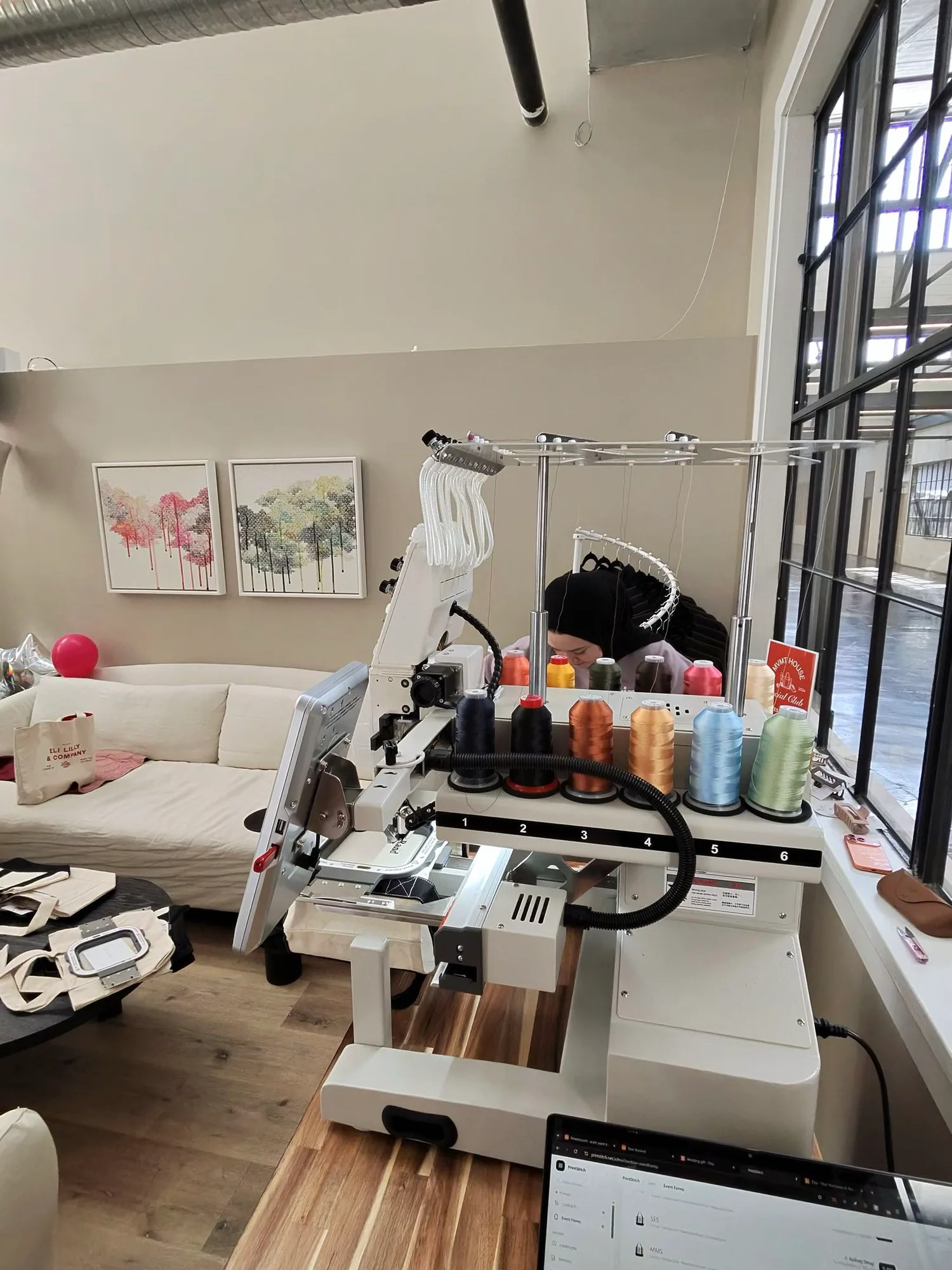 A PrintStitch embroidery machine at the MVMT House event, thread cones on the rack above it and canvas totes on the table